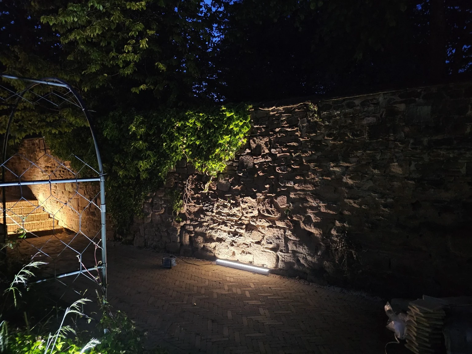 Warm architectural lighting washing textured stonework by garden arch
